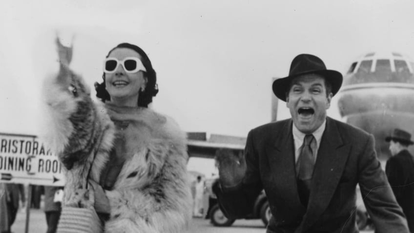 Vivien Leigh smiling and wear sunglasses as Laurence Olivier wearing a hat yells in excitement