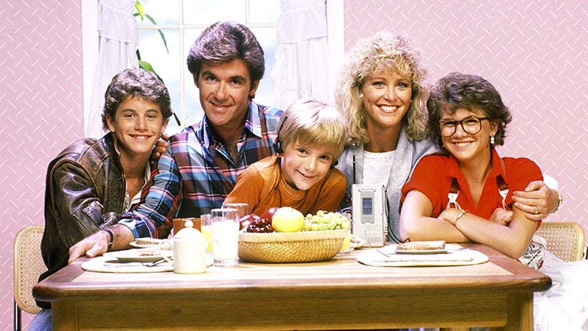 Kirk Cameron, Alan Thicke, Jeremy Miller, Joanna Kerns and Tracey Gold sit at a dinner table and smile for a family photo on the set of 'Growing Pains'