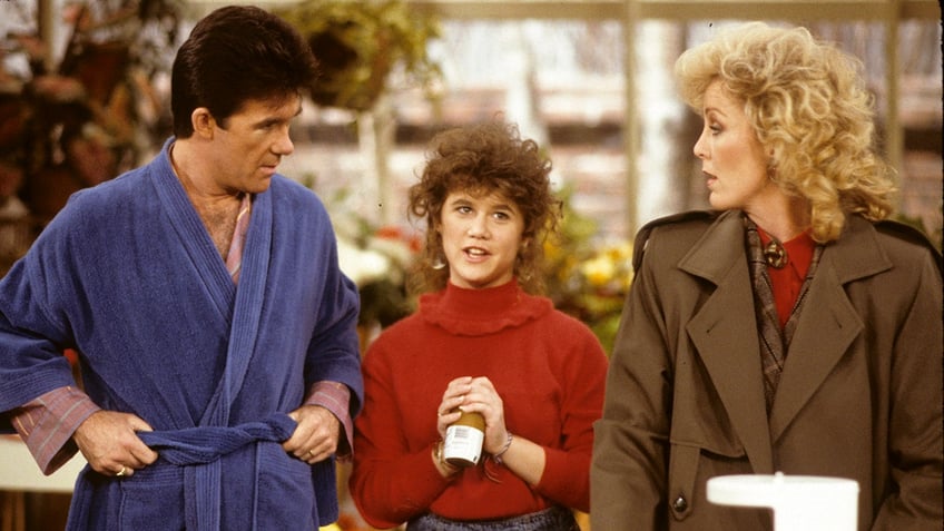 Alan Thicke in a blue bathrobe looks down at TV daughter Tracey Gold in a red sweater next to TV wife Joanna Kerns in a brown trench coat in a scene from "Growing Pains"