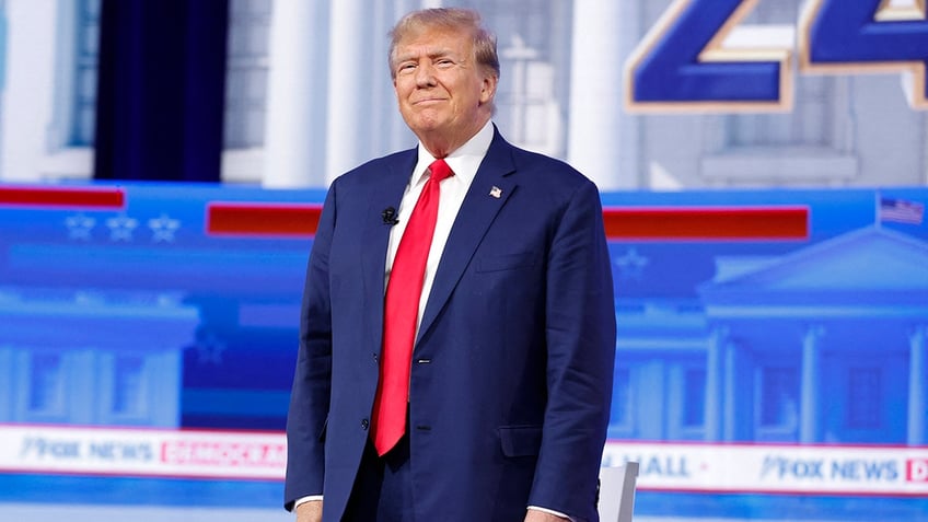 Trump smiling mouth closed, standing, hands by side, wearing a navy suit with a bright red tie