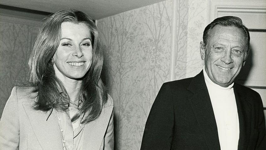 Stefanie Powers and William Holden smiling while walking next to each other.