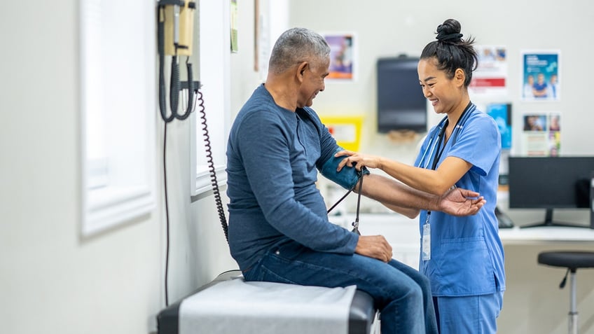 A man sits up on an exam table as his nurse takes his blood pressure