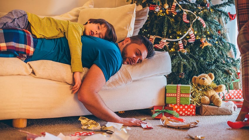 Father asleep on a sofa on Christmas Day, while his son lies asleep on his back