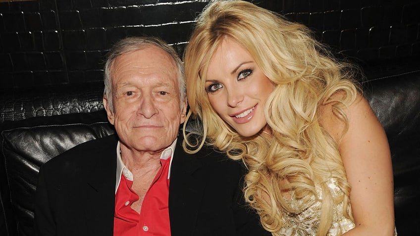 Hugh Hefner wearing a black blazer and a red shirt with Crystal Hefner leaning on him