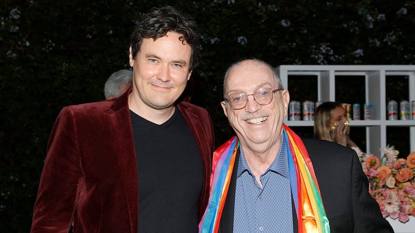 Gabriel Olds in a red velvet jacket soft smiles next to Steve Pieters in a blue shirt, black jacket, and rainbow scarf at a screening for "The Eyes of Tammy Faye"