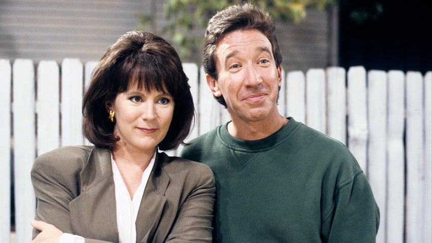 Patricia RIchardson as Jill Taylor in a brown blazer stands next to Tim Allen as Tim Taylor in a green sweater on "Home Improvement"