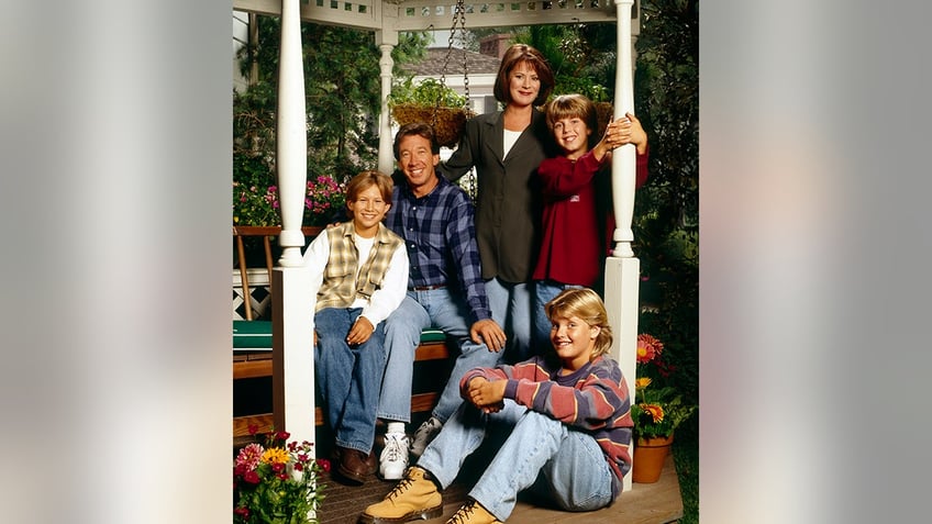 The cast of "Home Improvement" sitting on a bench hanging on to pillars outside the home