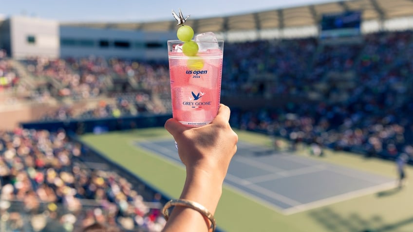 A hand holds the Honey Deuce cocktail into the air with the U.S. Open court in the background.