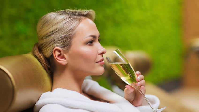 people, beauty, lifestyle, holidays and relaxation concept - beautiful young woman in white bath robe lying on chaise-longue and drinking champagne at spa