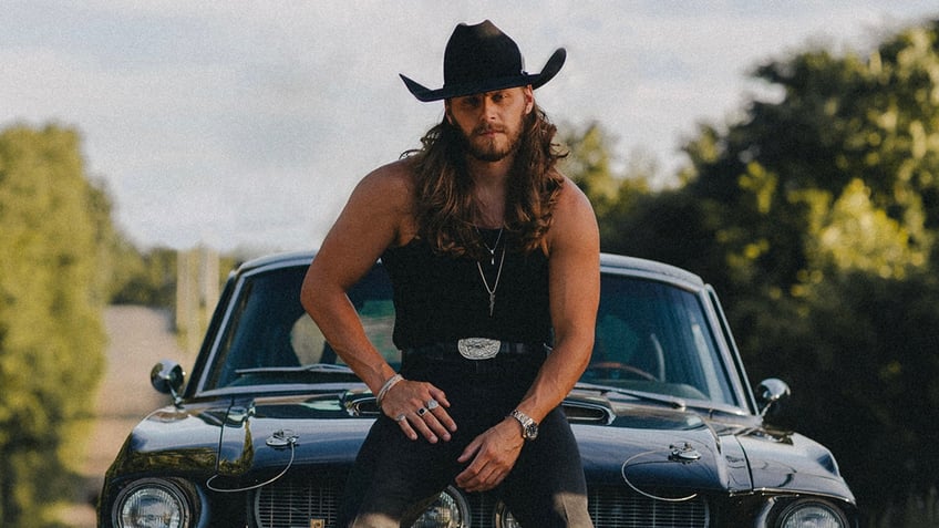 Warren Zeiders wearing all black and a black cowboy hat sitting on a car