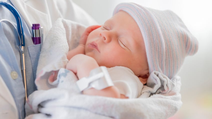 A Caucasian female infant is asleep in the arms of a pediatrician. The baby is swaddled in a blanket and is wearing a warm cap.