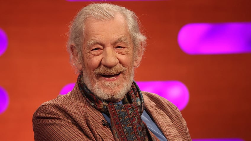 Ian McKellen in a burnt orange suit smiles on stage 