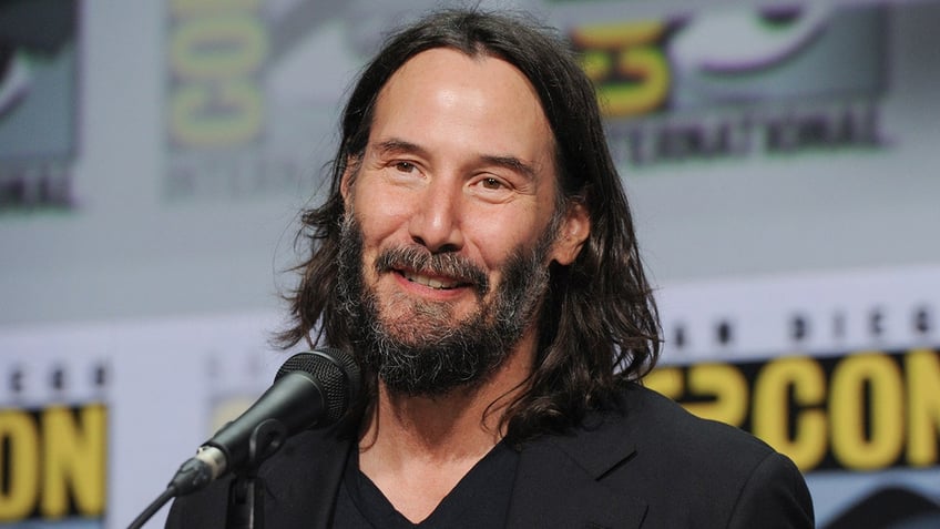 Keanu Reeves in a black shirt smiles as he speaks into a microphone at a Comic Con panel