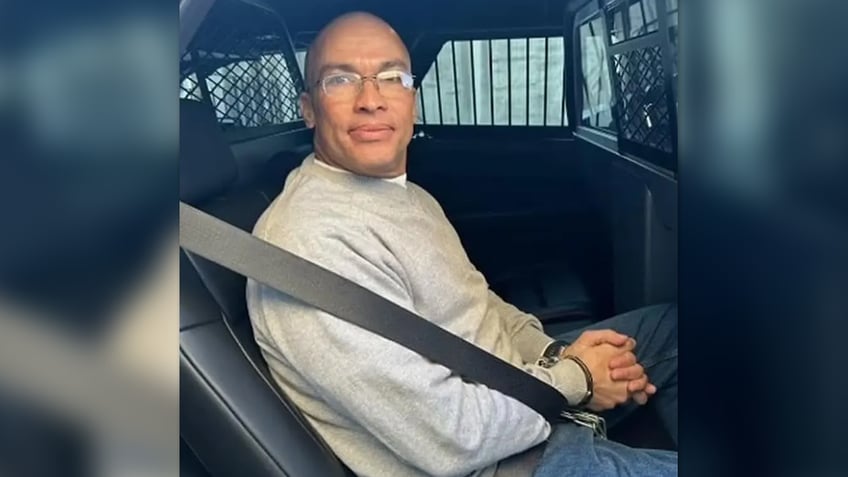 Cesar Polanco wearing jeans, a gray sweatshirt and glassses while handcuffed in the back of a law enforcement vehicle