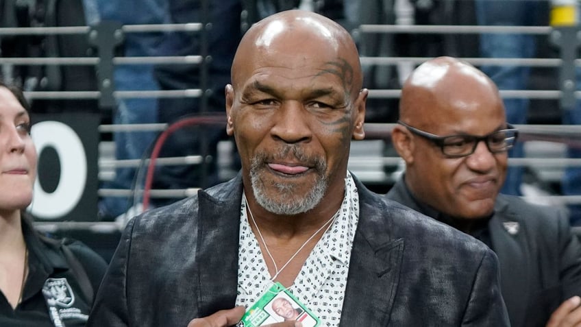 Former heavyweight boxing champion Mike Tyson wears a dark jacket and white shirt as he stands on the field before an NFL football game between the Las Vegas Raiders and the Pittsburgh Steelers in Las Vegas.