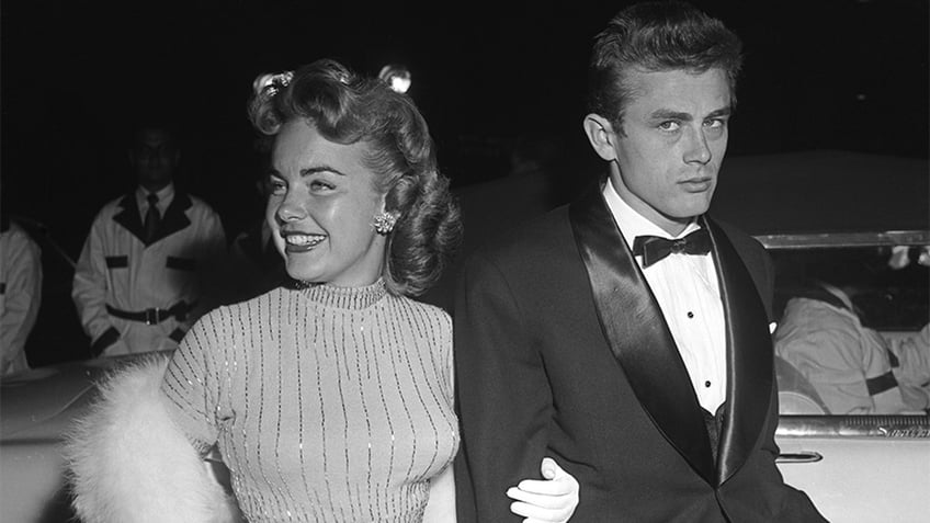 James Dean looking annoyed in a tux with a smiling woman holding onto his arm.