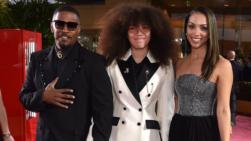 Jamie Foxx in a black suit poses with daughters Anelise and Corinne in a white blazer and black dress respectively at the Golden Globes