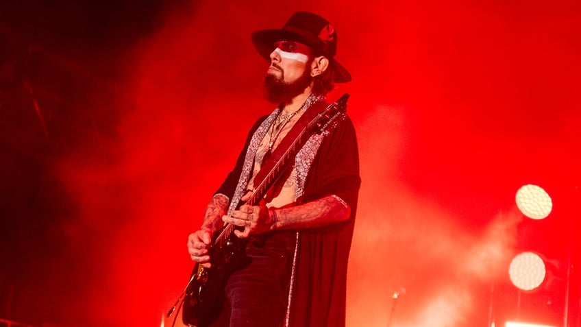 Dave Navarro in a black shirt plays guitar on stage with a red hue to the photo