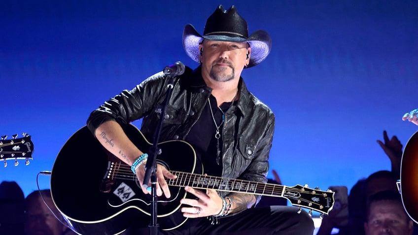 Jason Aldean in a black jacket and hat strums his black guitar on stage