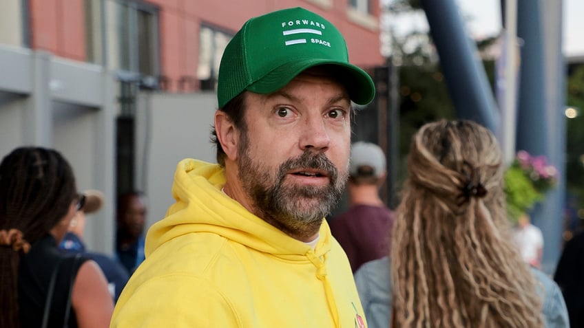 Jason Sudeikis wears a bright yellow sweatshirt and green cap outside of the US Open, looking a little startled by the camera