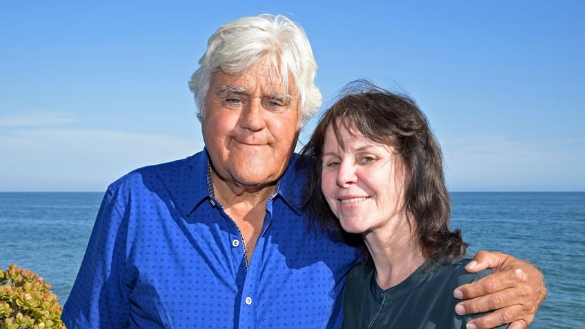 Jay Leno in a royal blue button down wraps his arm around wife Mavis in a dark teal shirt