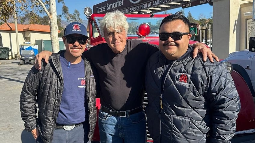 Jay Leno in a black shirt and jeans wraps his arms around two firefighters