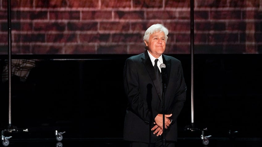 Jay Leno standing on stage in front of a brick wall