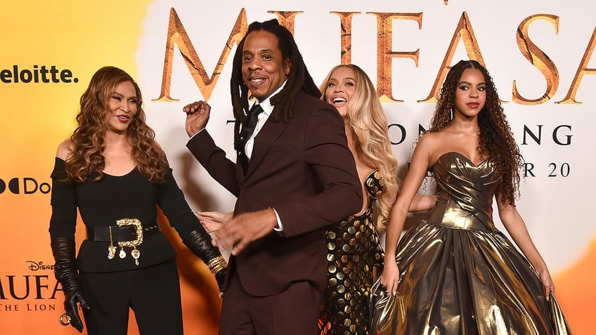 Jay-Z in a maroon suit smiles as he walks in front of his wife Beyoncé in a gold dress who laughs and daughter Blue Ivy in a gold dress and mother-in-law Tina Knowles in a black pantsuit