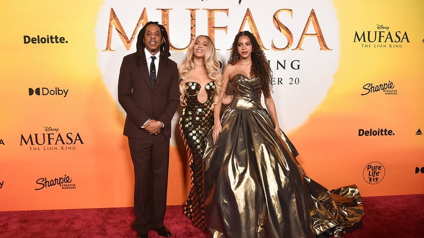 Jay-Z in a maroon suit stands with his hands beneath his waist next to wife Beyoncé in a gold plunging gown and daughter Blue Ivy in a strapless gold gown at 'Mufasa: The Lion King' premiere