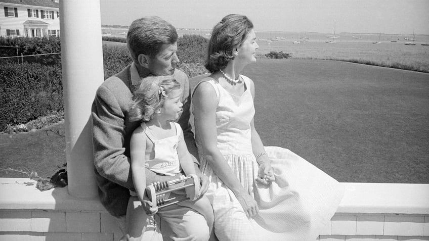 John F. Kennedy has Caroline on his lap sitting and looking out on the Cape with Jackie