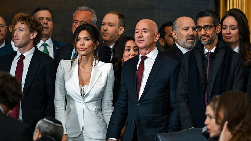 Mark Zuckerberg in a dark suit and red tie stands next to Lauren Sánchez in a white jacket next to Jeff Bezos in a dark suit