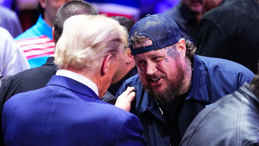 Jelly Roll beams wearing a backwards navy trucker hat and jacket while meeting Donald Trump (back to camera) at the UFC 309 event