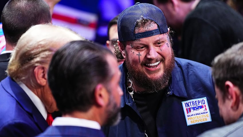 Jelly Roll smiles while speaking with (partially obscured) Donald Trump, Donald Trump Jr. and Elon Musk at the UFC 309 event