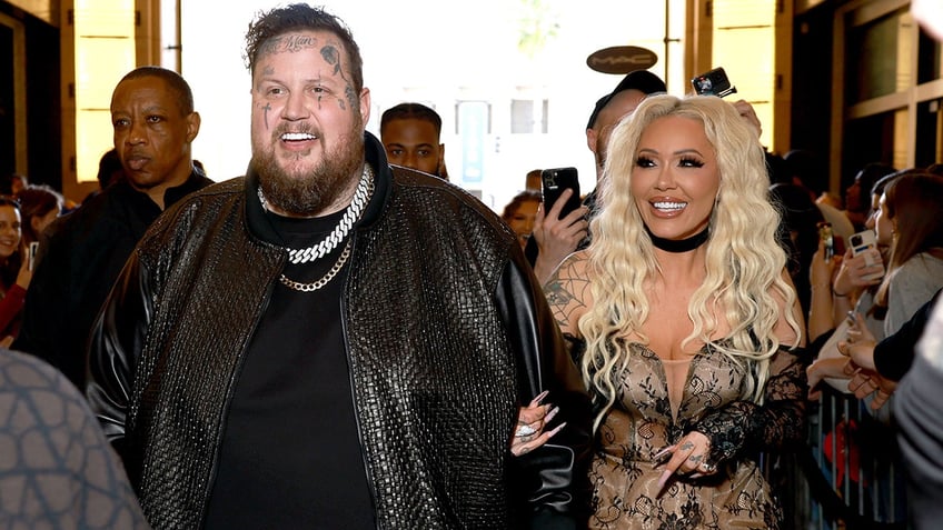 Jelly Roll in a black leather jacket walks into the iHeartRadio Music Awards with his wife Bunnie Xo in a lace black outfit