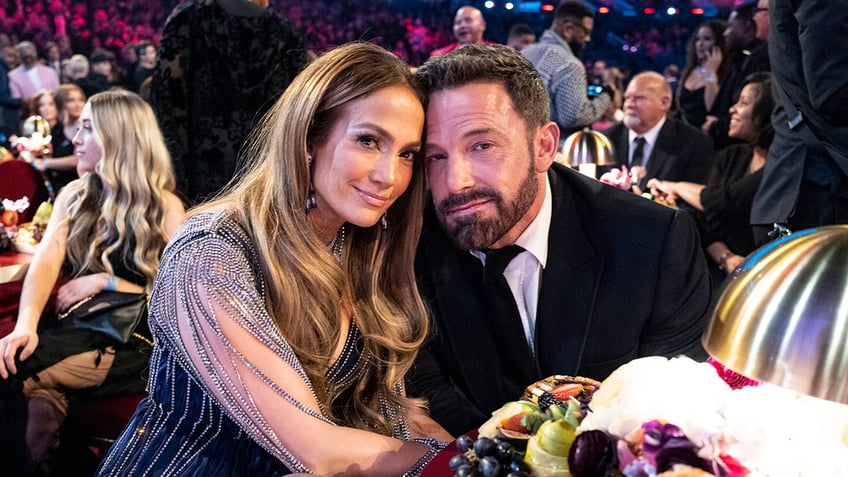 Jennifer Lopez iin a blue dress with sheer sleeves smiles softly with husband Ben Affleck in a black suit at the Grammys