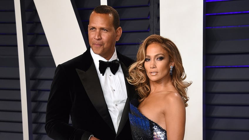 Alex Rodriguez in a black tuxedo looking to his right and Jennifer Lopez in a blue one shoulder dress looking to her right