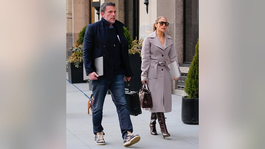 Ben Affleck in a peacoat and jeans walks with his computer in one arm and a briefcase in the other with Jennifer Lopez in a grey coat