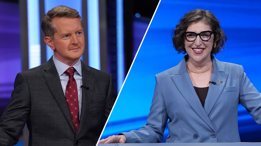 Ken Jennings in a dark suit and red tie looks to contestants while on Jeopardy! split Mayim Bialik in a blue jacket behind a podium on "Jeopardy!"