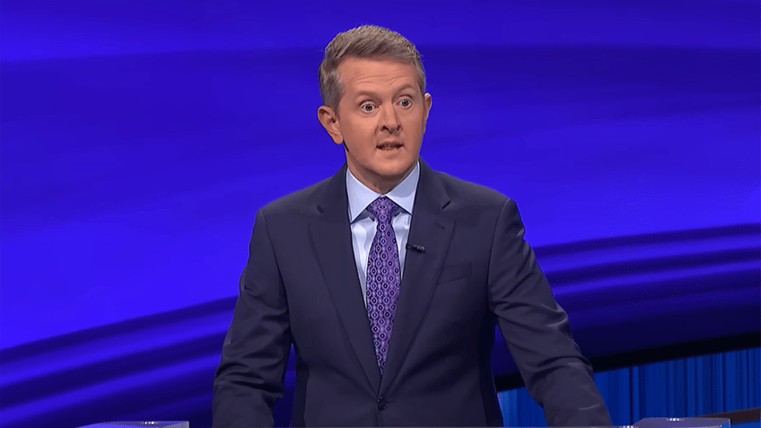 Ken Jennings in a navy suit with a light blue shirt and purple suit on the set of 'Jeopardy!'