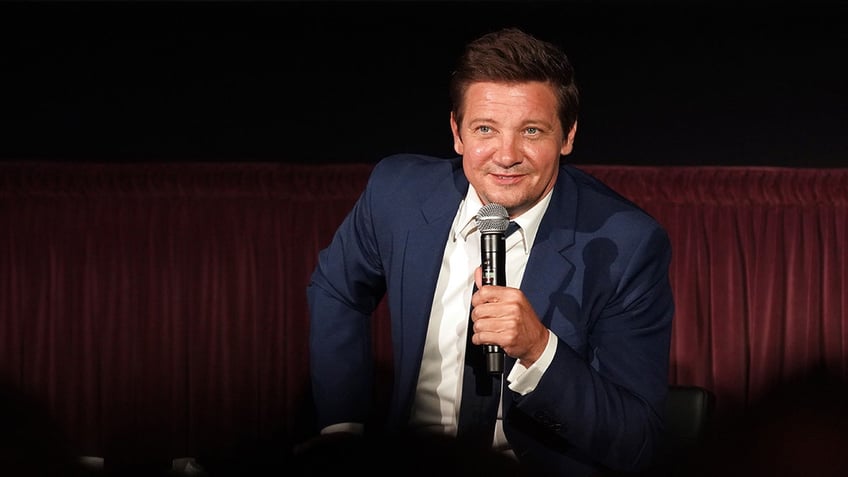 Jeremy Renner holding a mic wearing a navy suit and smiling