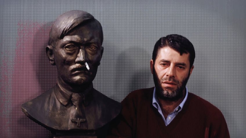 Jerry Lewis wearing a dark sweater and a light blue shirt looking serious as he poses next to a statue that has a cigarette up its nostril.