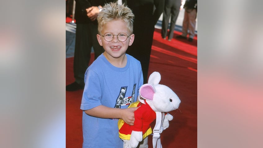 Jonathan Lipnicki in a blue t-shirt on the carpet holding a 'Stuart Little' toy