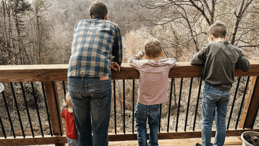 Derick in a plaid shirt looks over a deck with sons Israel, 9, Samuel, 6, and Fredrick, 1