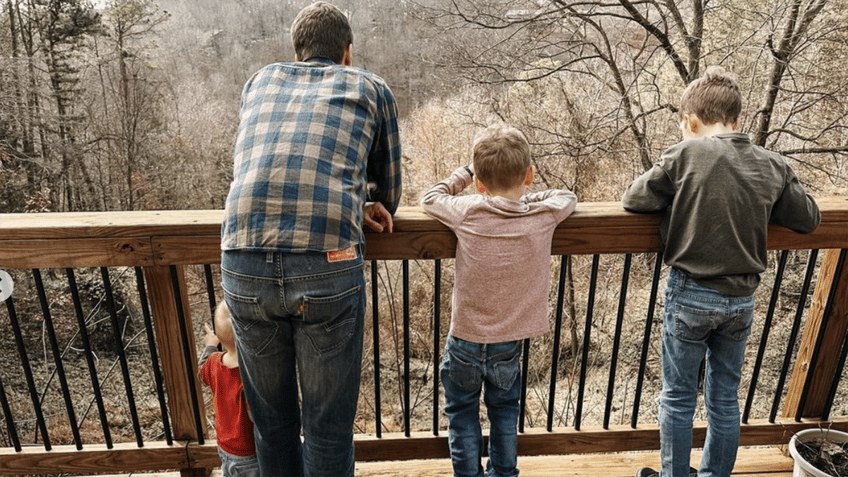 Derick in a plaid shirt looks over a deck with sons Israel, 9, Samuel, 6, and Fredrick, 1