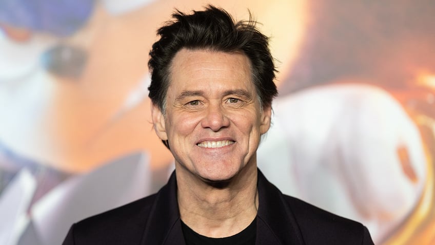 Jim Carrey smiles directly at the camera on the carpet, wearing a black shirt and jacket in London