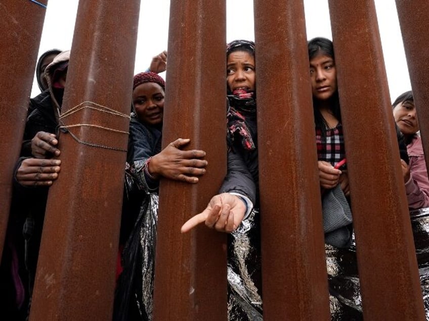 Migrants reach through a border wall for clothing handed out by volunteers, as they wait b