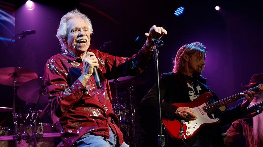 Joe Bonsall in a red shirt and jeans holds on to a microphone stand and sings with The Oak Ridge Boys