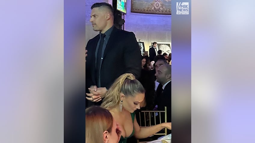 Joe Manganiello in a black suit speaks to someone as his girlfriend Caitlin sits at the table below him