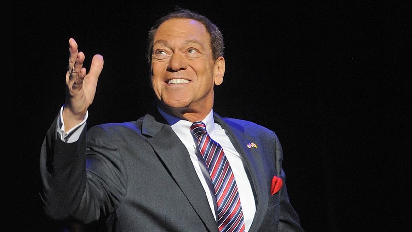 Joe Piscopo looking up in a dark grey suit smiling wearing with a colorful striped tie.