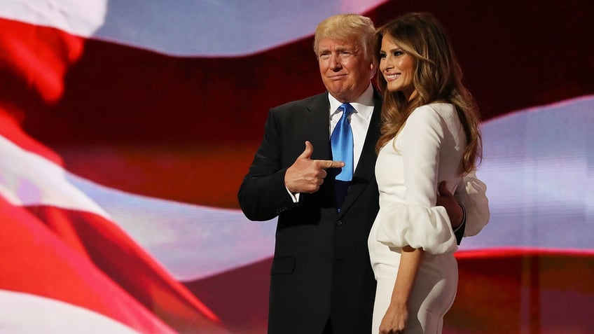 Donald Trump wearing a dark suit, a white shirt and a light blue tie pointing at his wife Melania Trump who is wearing a white dress